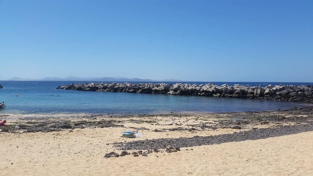 Sandy beach with scattered dark rocks in the foreground, calm clear blue water, and a breakwater made of large concrete blocks. Distant hazy mountains are visible on the horizon under a clear blue sky. Beach towels and bags lie on the sand.