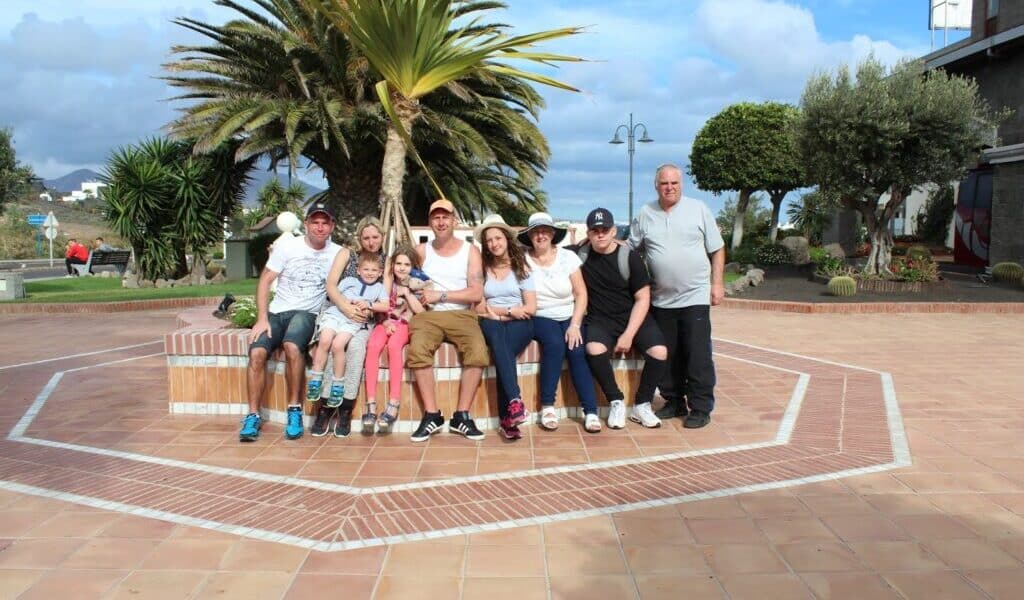 A group of nine people, including adults and children, sit closely together on a brick bench in a sunny outdoor plaza surrounded by palm trees and landscaping, smiling at the camera. The sky is partly cloudy, and the ground is tiled.