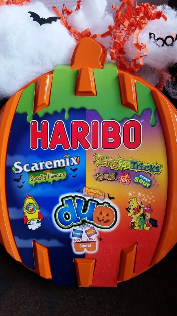 A plastic pumpkin-shaped box features Halloween-themed HARIBO sweets. The lid is colourful with the HARIBO logo and labels for Scaremix, Tangfastricks, and Duo sweets. Faux spider webs, bats, and orange decorations are in the background.
