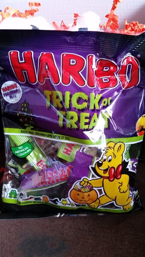 A bag of Haribo Trick or Treat sweets features a yellow cartoon bear holding a pumpkin basket. The bag is purple with Halloween-themed graphics, and individual sweet packets are visible inside. The text says, “Kids and grown-ups love it so.”.