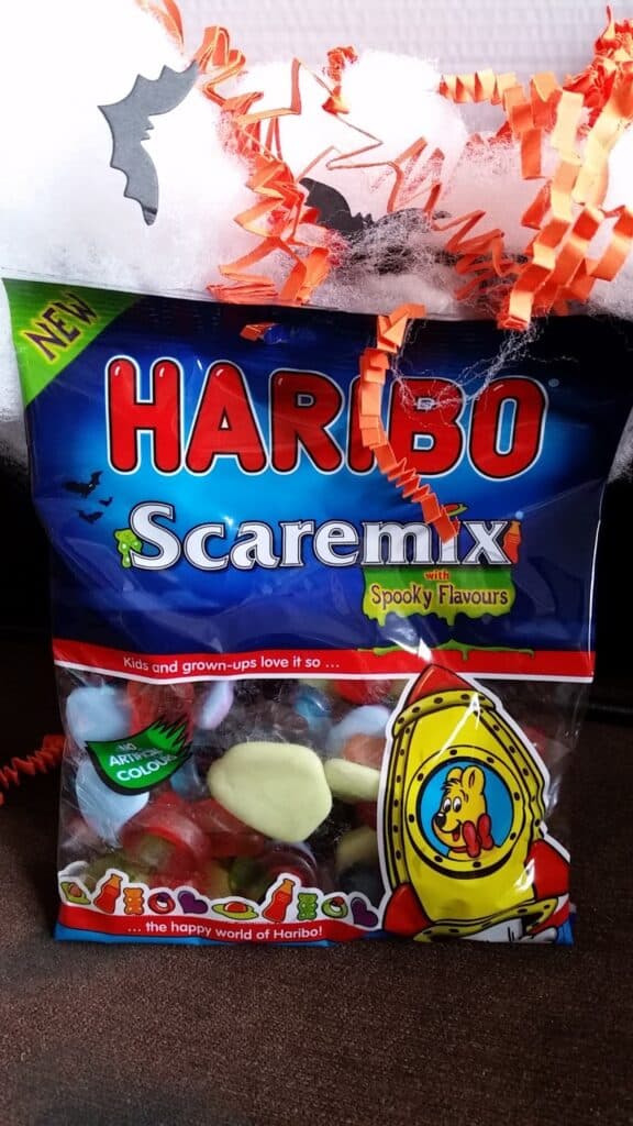 A bag of Haribo Scaremix gummy sweets sits on a surface, decorated for Hallowe’en with white spider webbing, an orange paper coil, and a black paper bat. The packet is dark blue, advertising “spooky flavours” and “vibe-o-licious” sweets.