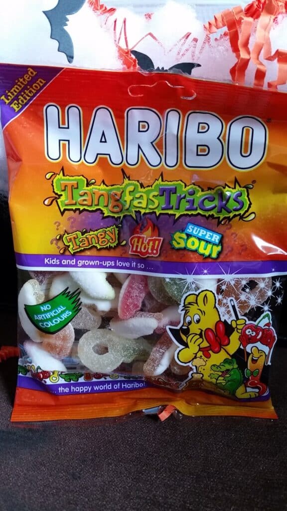 A bag of HARIBO TangfasTricks limited edition gummy sweets, featuring flavours labelled as tangy, hot, and super sour. The packaging is colourful, with cartoon gummy bears, and highlights "No Artificial Colours" on the front. Assorted gummies are visible inside.