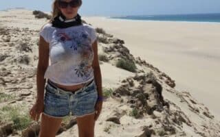 A woman wearing sunglasses, a white T-shirt, denim shorts, and sandals stands on sandy dunes under a clear blue sky. The sea is visible in the background, and grass grows sparsely among the sand.