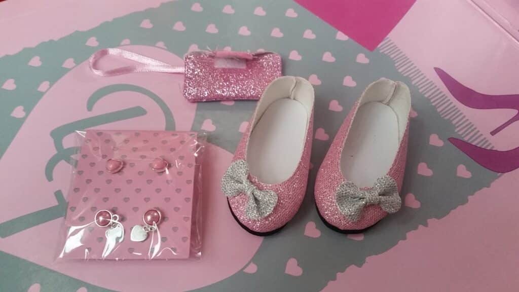 A pair of sparkly pink doll shoes with silver bows, a matching glittery pink handbag, and a set of pink and silver earrings and charms arranged on a pastel pink and blue heart-patterned background.