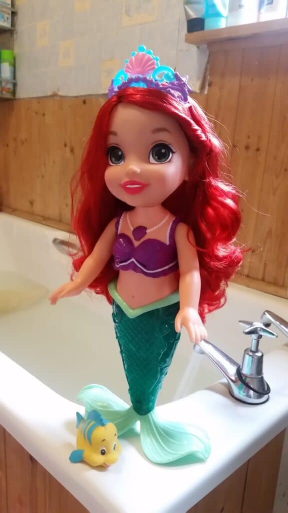 A doll of Ariel from Disney’s “The Little Mermaid” with red hair, a purple seashell top, and green mermaid tail stands by a bath tap. A small yellow and blue Flounder toy is beside her. The background shows wooden panelling and bathroom items.