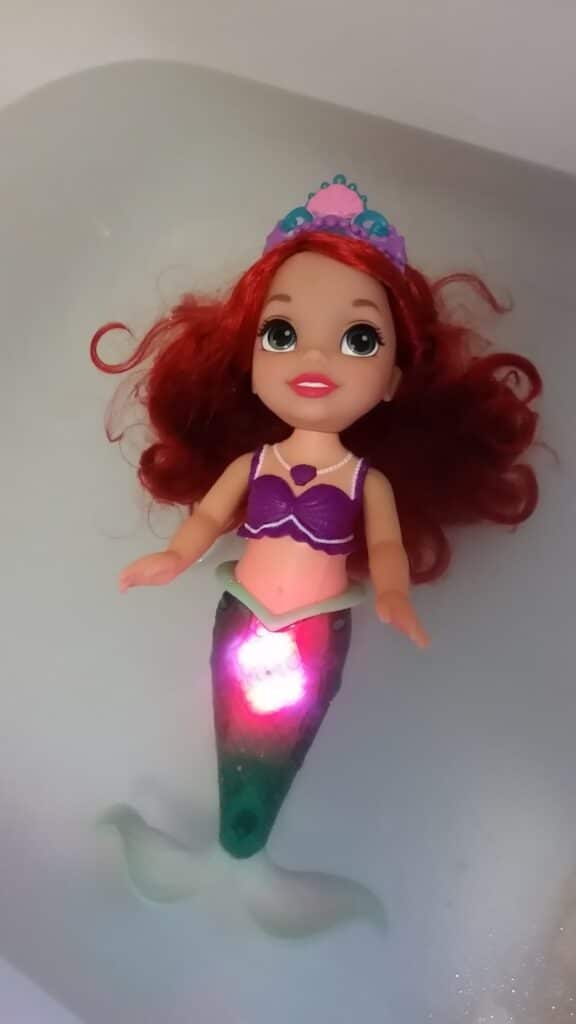 A plastic mermaid doll with long red hair, a purple shell top, and a green tail floats in water. She wears a colourful tiara and has a light glowing from her tail, illuminating part of her body. The background is a plain bath.
