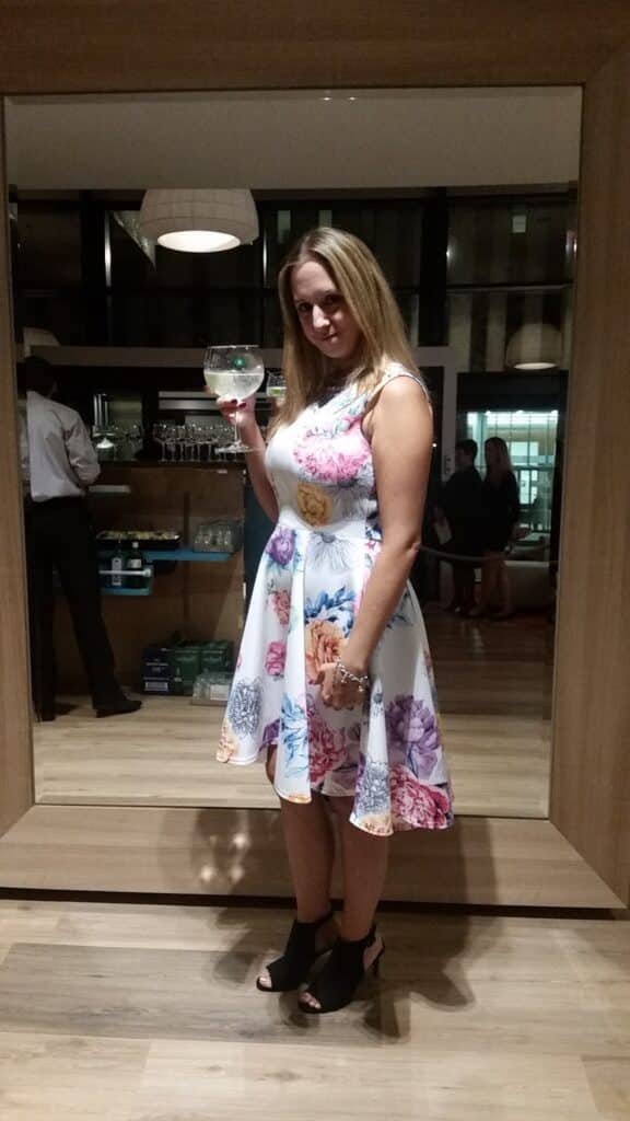 A woman in a white, sleeveless floral dress and black open-toe heels holds a large glass of clear drink. She stands in front of a large mirror in a stylish indoor setting with wooden floors, shelves, and people in the background.