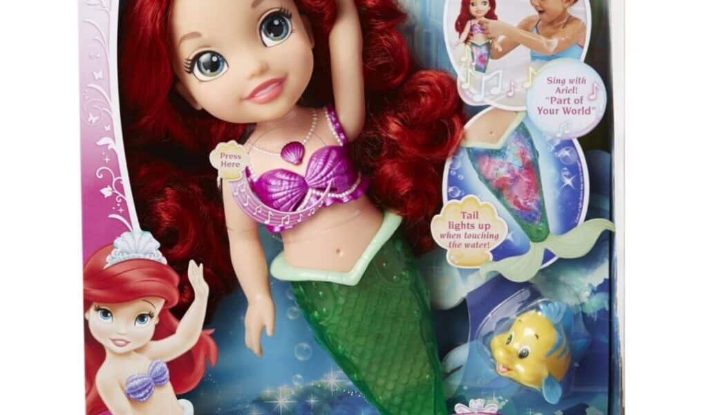 A boxed Disney Princess "Colours of the Sea Ariel" doll with red hair, wearing a purple seashell top and green mermaid tail. The packaging features images of Ariel, a child playing, Flounder, and highlights that Ariel's tail lights up and plays music.