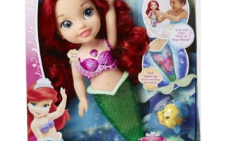 A boxed Disney Princess "Colours of the Sea Ariel" doll with red hair, wearing a purple seashell top and green mermaid tail. The packaging features images of Ariel, a child playing, Flounder, and highlights that Ariel's tail lights up and plays music.