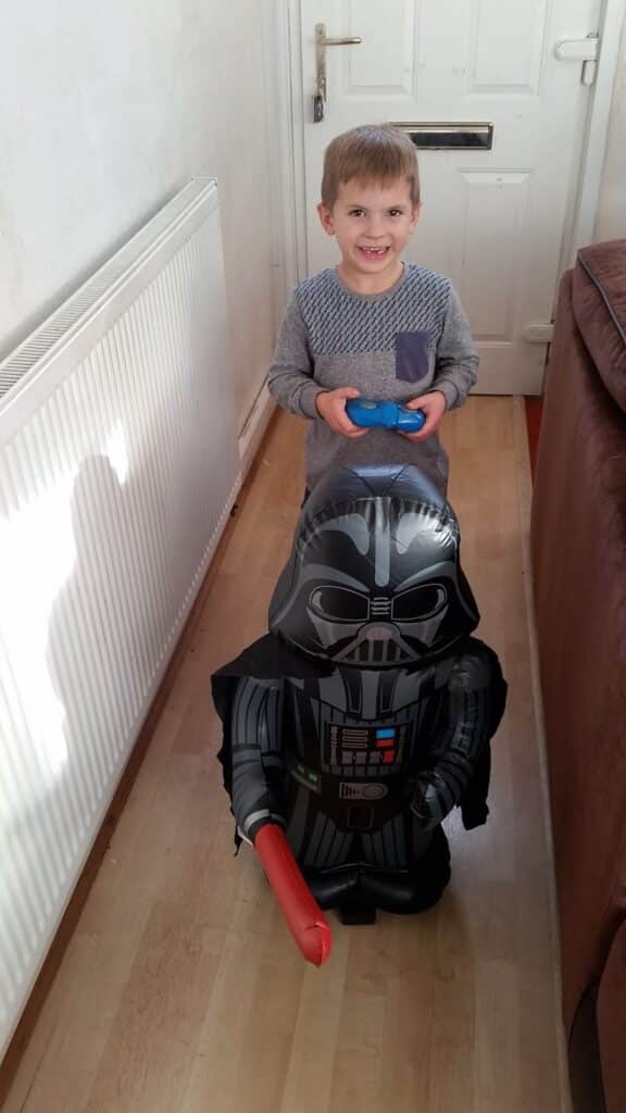 A smiling young boy stands indoors on a wooden floor, holding a blue object. In front of him is a large inflatable Darth Vader figure holding a red lightsabre. Sunlight casts shadows on the white radiator and wall to the left.