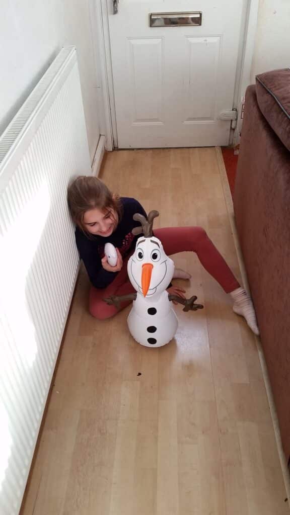 A young girl with light brown hair in a ponytail, wearing a navy jumper and pink leggings, sits on a light wooden floor near a radiator. She hugs an inflatable Olaf snowman toy from Disney’s “Frozen” and smiles up at the camera in a narrow corridor.