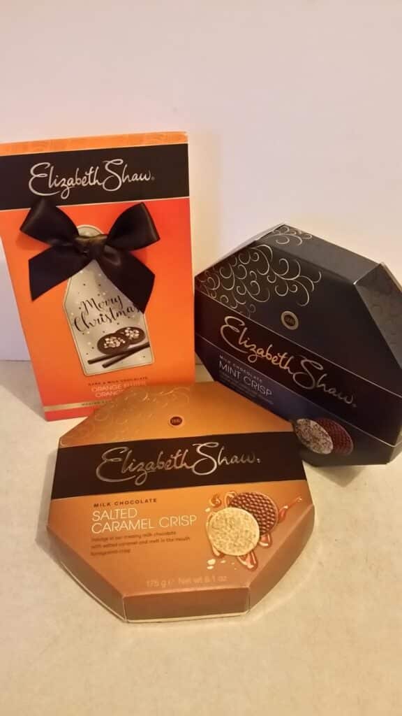Three boxes of Elizabeth Shaw chocolates are shown: one orange box with a festive tag and black ribbon, one hexagonal orange box labelled "Milk Chocolate Salted Caramel Crisp," and one hexagonal dark box labelled "Mint Crisp.
