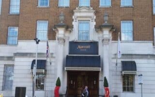 A person stands near the entrance of a large brick and white stone hotel building. Above the door is a black awning with “Marriott Hotel” written on it. Two flags and trimmed bushes flank the entrance, and multiple windows line the upper floors.