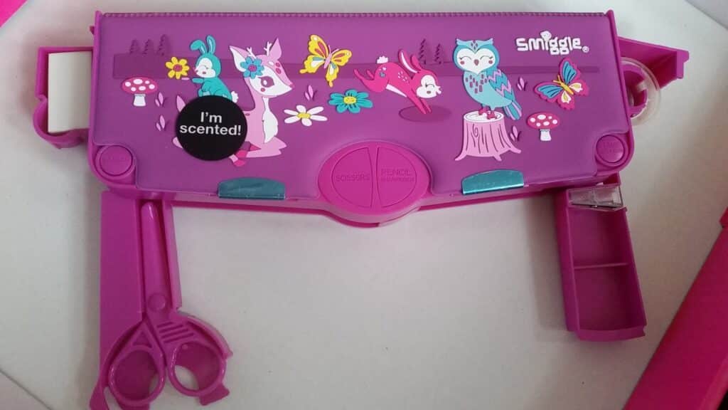 A pink Smiggle pencil case decorated with cute animal illustrations, butterflies, and mushrooms. It has compartments open with visible scissors, tape, and rubber. A black sticker reads, "I'm scented!" The case is brightly coloured and playful.