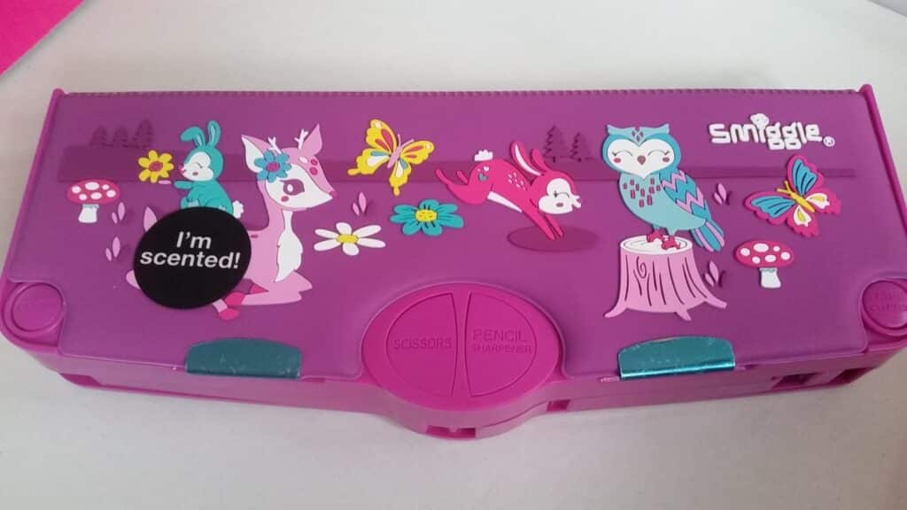 A purple Smiggle pencil case decorated with cute illustrations of a rabbit, deer, fox, owl, butterflies, flowers, and mushrooms. A black sticker says “I’m scented!” The case has two green clasps and labelled sections for scissors and a pencil sharpener.