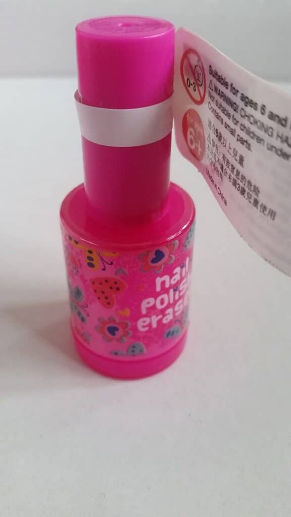 A small, bright pink bottle with a label showing cartoon ladybirds and hearts. The label reads "nail varnish remover" in white text. A white tag attached to the cap says “Suitable for ages 6+.” The bottle is standing on a plain white surface.