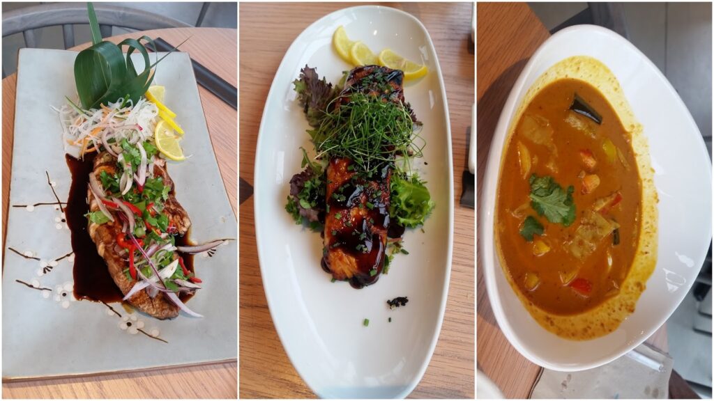 Three dishes on wooden tables: left, a rectangular plate with grilled meat topped with herbs, sliced red onion, and a lemon wedge; centre, an oval plate with glazed grilled pieces and greens; right, a white bowl of yellow curry with vegetables and coriander.