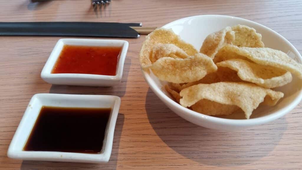 A white bowl filled with prawn crackers sits on a light wooden table. Next to it are two small square dishes: one with a dark soy sauce and one with a bright orange sweet chilli sauce. Chopsticks and a fork are in the background.