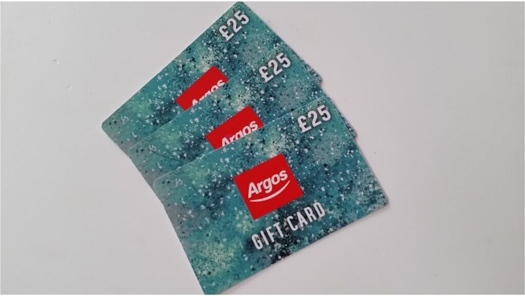 Three Argos gift cards, each valued at £25, are fanned out on a white surface. The cards have a speckled blue-green background and display the Argos logo in red, with the word "GIFT CARD" written in white underneath.
