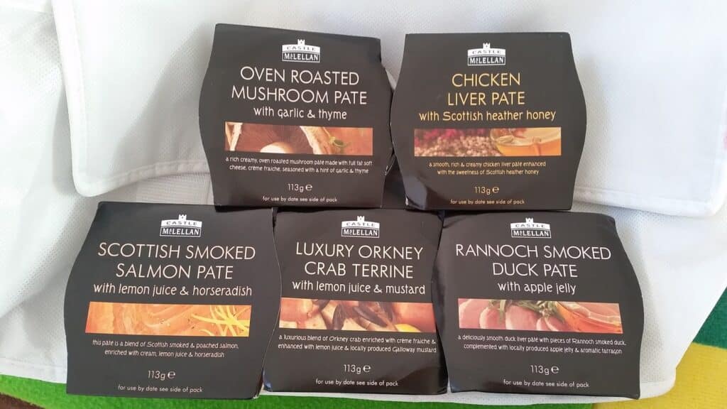 Five square packs of Castle MacLellan pâté are arranged in two rows. Flavours include: Oven Roasted Mushroom, Chicken Liver, Scottish Smoked Salmon, Luxury Orkney Crab Terrine, and Rannoch Smoked Duck. Each pack shows ingredients and weighs 113g.