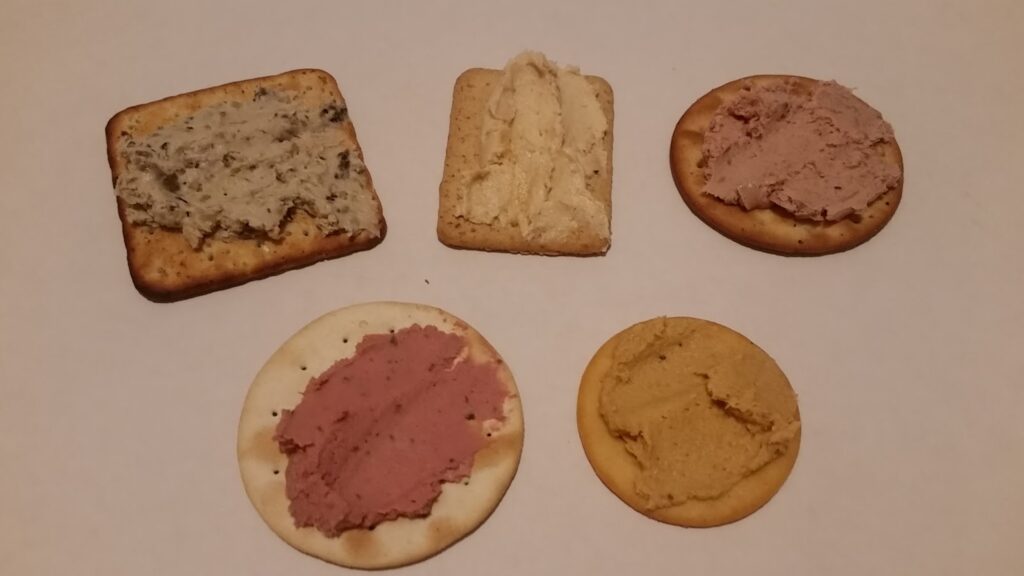 Five biscuits on a white surface, each topped with a different spread. The spreads vary in colour and texture: grey with herbs, beige, pinkish-brown, and yellow. The biscuits are a mix of round and rectangular shapes.