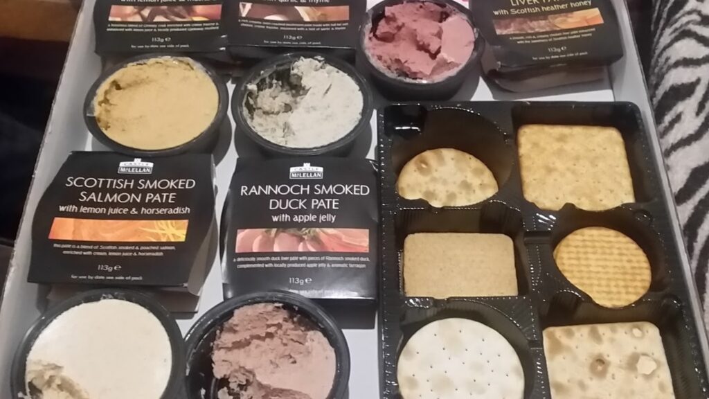 A selection box contains three open pâtés—Scottish smoked salmon, Rannoch smoked duck, and venison—each in black containers, along with an assortment of round and square biscuits in separate sections of the tray. The pâté looks creamy and spreadable.