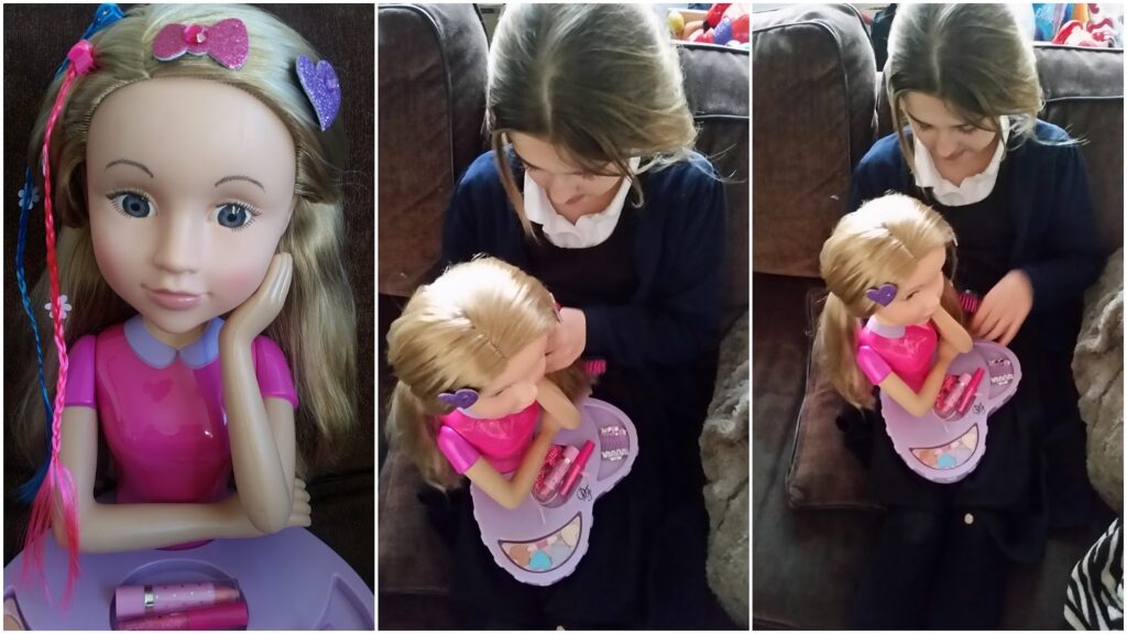 A young girl sits on a sofa playing with a large styling head doll. The doll, with blonde hair and bright eyes, is decorated with colourful hair clips and plaits. The girl, dressed in a dark dress, smiles whilst brushing and styling the doll’s hair.