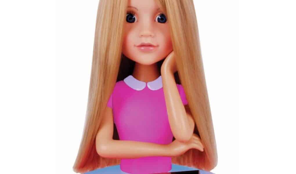 A blonde styling doll head with long straight hair, blue eyes, and a hand resting on its cheek. The doll wears a pink top with a white collar. In front, on a purple heart-shaped tray, are hair accessories and toy make-up items.