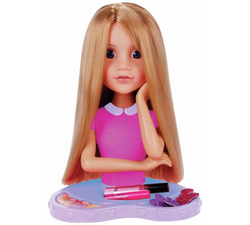 A blonde doll styling head with long straight hair, blue eyes, and a pink dress top with a white collar rests its chin on one hand. The doll sits on a purple stand with hearts and is surrounded by hair accessories and lip gloss.