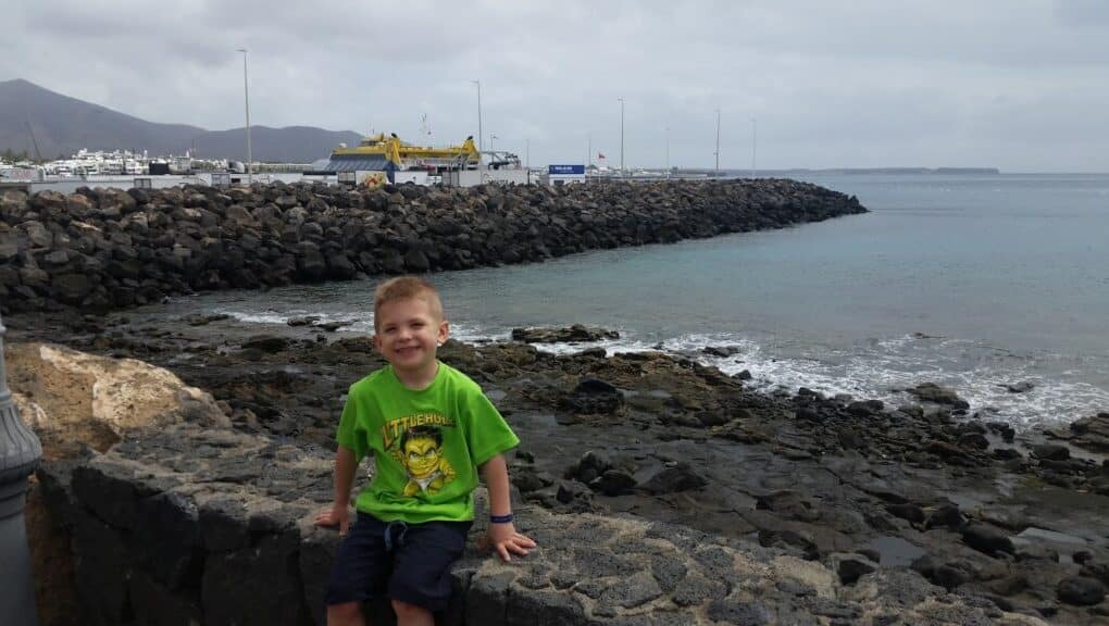 A young boy in a bright green T-shirt and navy shorts sits on a stone wall by the rocky shore, smiling at the camera. Behind him, calm sea waves touch dark rocks, and a harbour with cranes and boats is visible under a cloudy sky.
