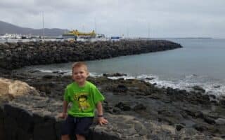 A young boy in a bright green T-shirt and navy shorts sits on a stone wall by the rocky shore, smiling at the camera. Behind him, calm sea waves touch dark rocks, and a harbour with cranes and boats is visible under a cloudy sky.