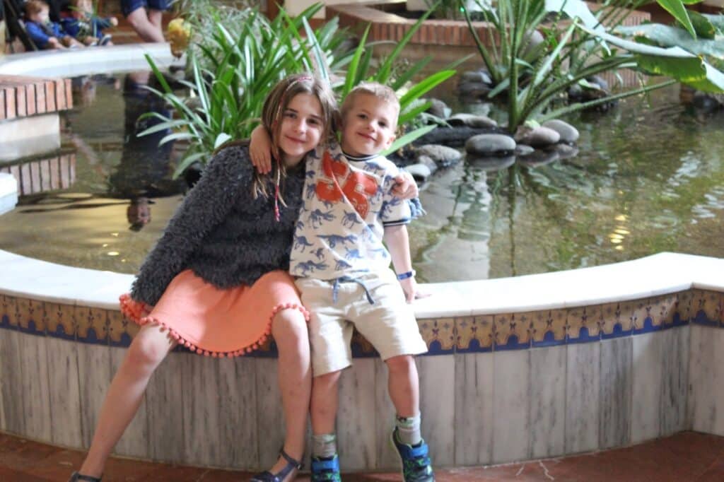 Two young children sit side by side on the edge of a round indoor fountain. The girl in a grey jumper and peach skirt hugs the boy, who wears a dinosaur-print shirt and beige shorts. They both smile, with green plants and water behind them.