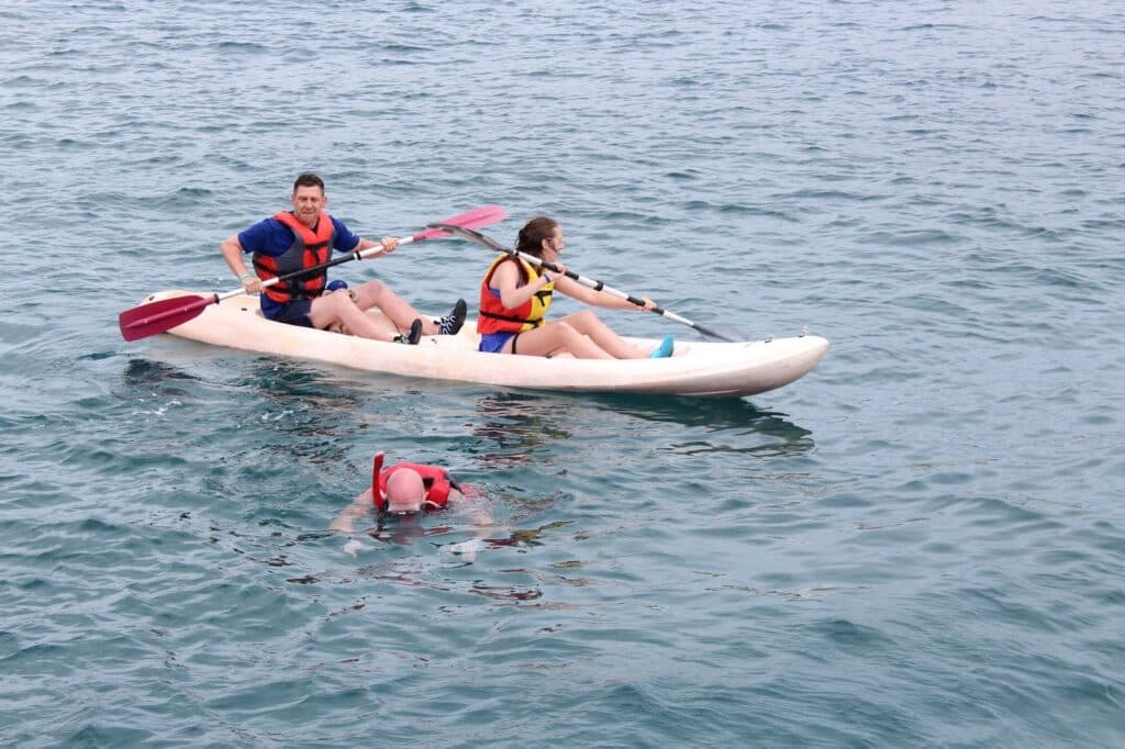 Two people in life jackets paddle a white kayak on calm blue water, while a third person wearing a red life jacket swims nearby. The kayakers look ahead as the swimmer, mostly submerged, moves beside the kayak.
