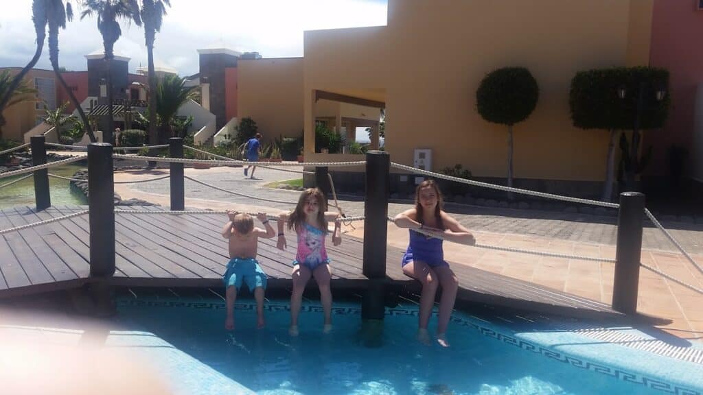 Three children sit on the edge of a wooden bridge over a pool, splashing their feet in the water. Two girls and one boy are in swimming costumes, with buildings, palm trees, and a person walking in the sunny background.
