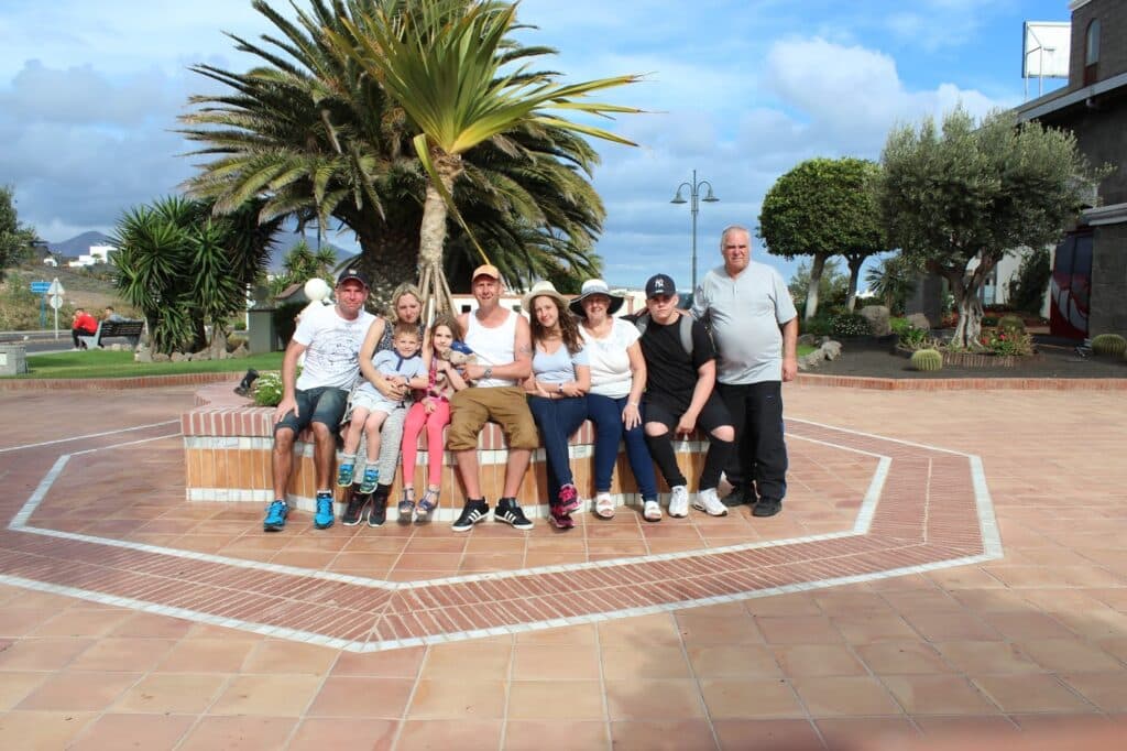 Eight people sit closely together on a brick bench in a sunny plaza with palm and other trees behind them. Most are casually dressed in T-shirts and shorts. The group includes adults and children, all smiling at the camera. The sky is partly cloudy.