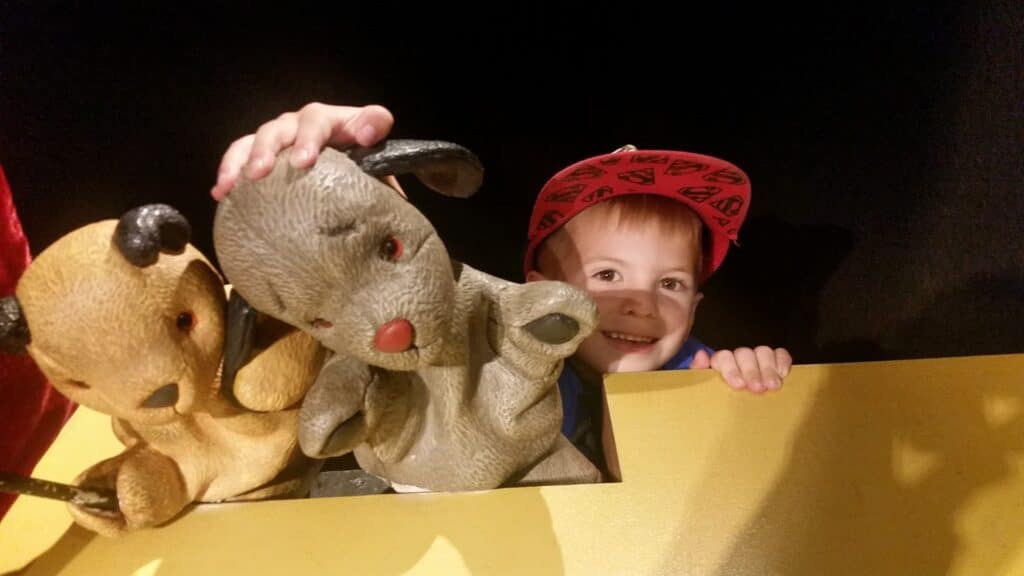 A smiling young child in a red cap is behind a yellow puppet stage, holding two puppet dogs, one yellow and one grey, both with floppy ears and red noses, set against a dark background.
