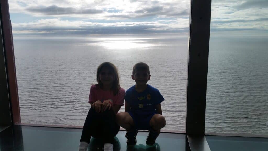 Two children, a girl in a pink shirt and a boy in a blue shirt, sit side by side on the floor in front of a large window overlooking a vast, calm body of water under a cloudy sky with sunlight reflecting on the surface.