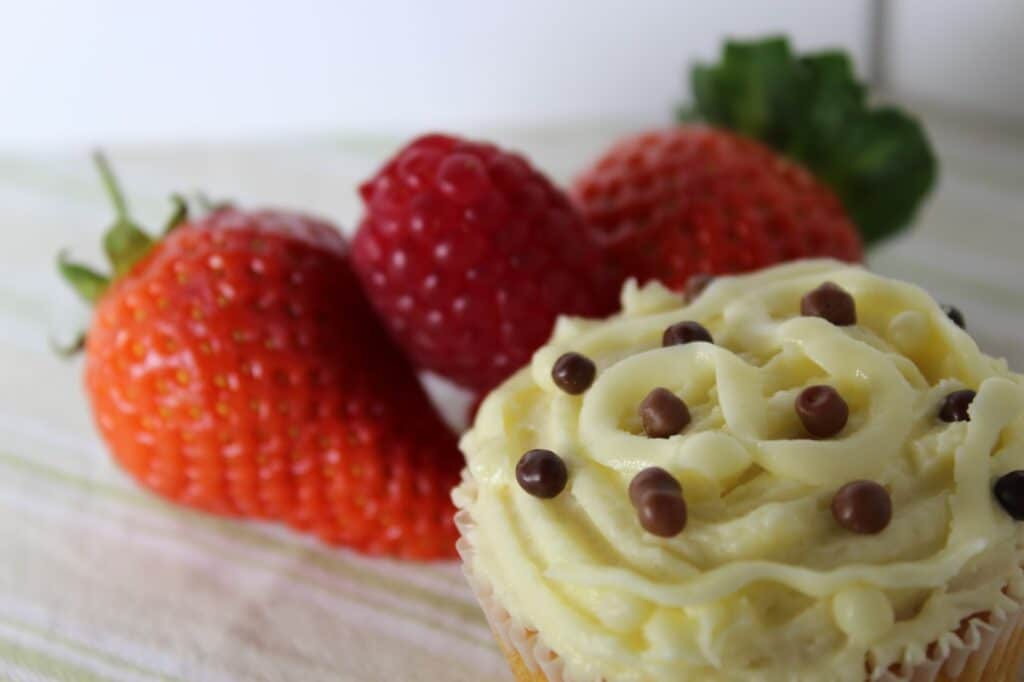 A close-up of a cupcake with creamy white icing and chocolate chips, with two strawberries and a raspberry in the blurred background on a light striped surface. The image has a soft, inviting look and emphasises the dessert and fruit.