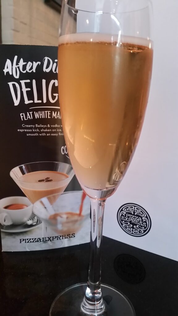 A champagne flute filled with a light golden sparkling drink stands on a dark surface. Behind it is a menu featuring a martini cocktail and the words “After Dinner Delight” and “PizzaExpress.” The logo of PizzaExpress is partly visible on the right.