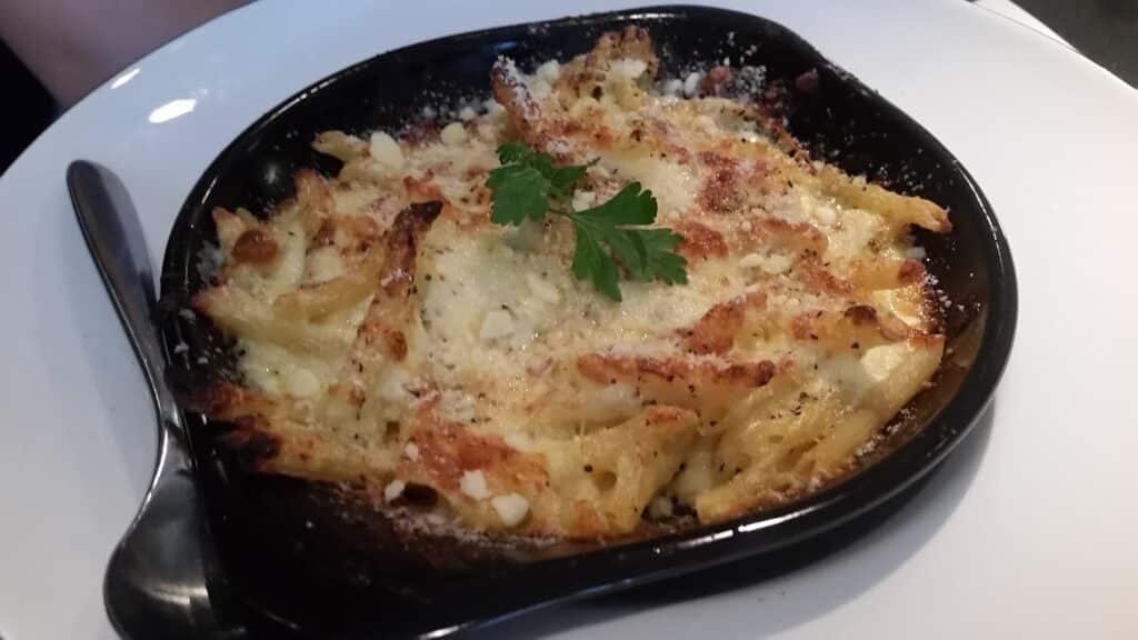 A black dish filled with baked penne pasta topped with melted cheese and herbs sits on a white plate. The pasta is golden brown with crispy edges, and a sprig of fresh parsley is placed in the centre. A black spoon rests beside the dish.
