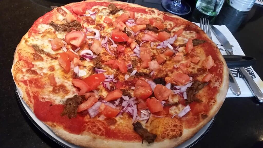 A large pizza with a golden-brown crust, topped with melted cheese, chunks of tomato, sliced red onion, pieces of beef, and tomato sauce, is on a round tray. Cutlery, a serviette, and a glass of water are visible in the background on a dark table.