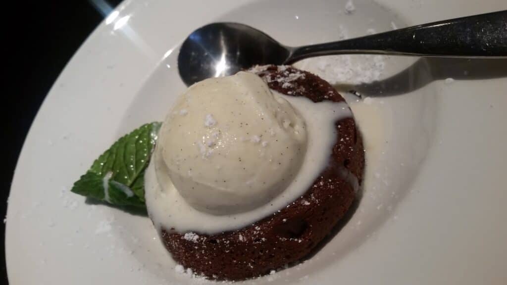 A round chocolate fondant is topped with a melting scoop of vanilla ice cream and garnished with a fresh mint leaf. Icing sugar is sprinkled on the white plate, and a spoon rests beside the pudding.