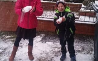 Two smiling children stand outside on a snowy pavement. The girl on the left wears a bright pink coat and hat, holding a snowball. The boy on the right, in a green and black coat with a rucksack, also holds a snowball. Cars and houses are visible in the background.