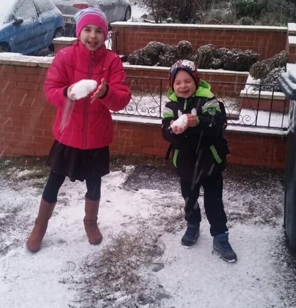 Two smiling children stand outside on a snowy pavement. The girl on the left wears a bright pink coat and hat, holding a snowball. The boy on the right, in a green and black coat with a rucksack, also holds a snowball. Cars and houses are visible in the background.