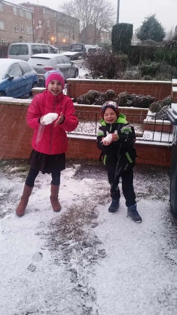 Two children, a girl in a pink coat and hat, and a boy in a black jacket with green accents, smile while holding snowballs outside. Snow covers the ground and parked cars. Houses and bushes are visible in the background on a snowy day.