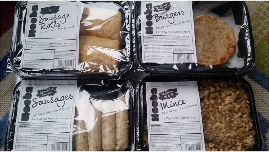 Four black plastic trays of packaged vegan meat alternatives are shown: sausage rolls (top left), burgers (top right), sausages (bottom left), and minced “meat” (bottom right). Each has a white paper label with product details and ingredients.