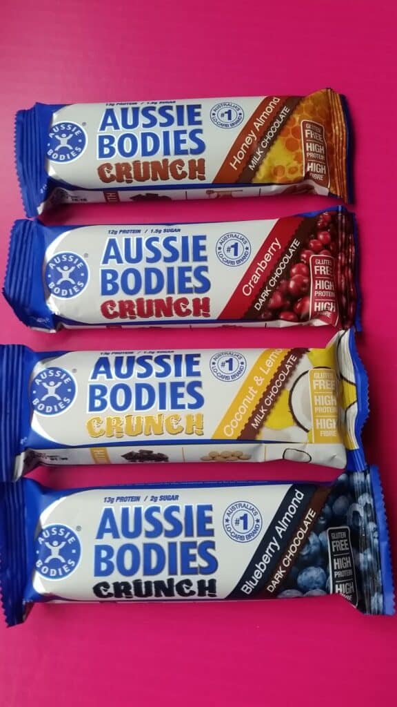 Four Aussie Bodies Crunch protein bars are stacked on a pink surface. Flavours from top to bottom: Honey Almond, Cranberry, Coconut & Lemon, and Blueberry Almond. Each bar features colourful wrappers with high protein, high fibre, and low sugar labels.