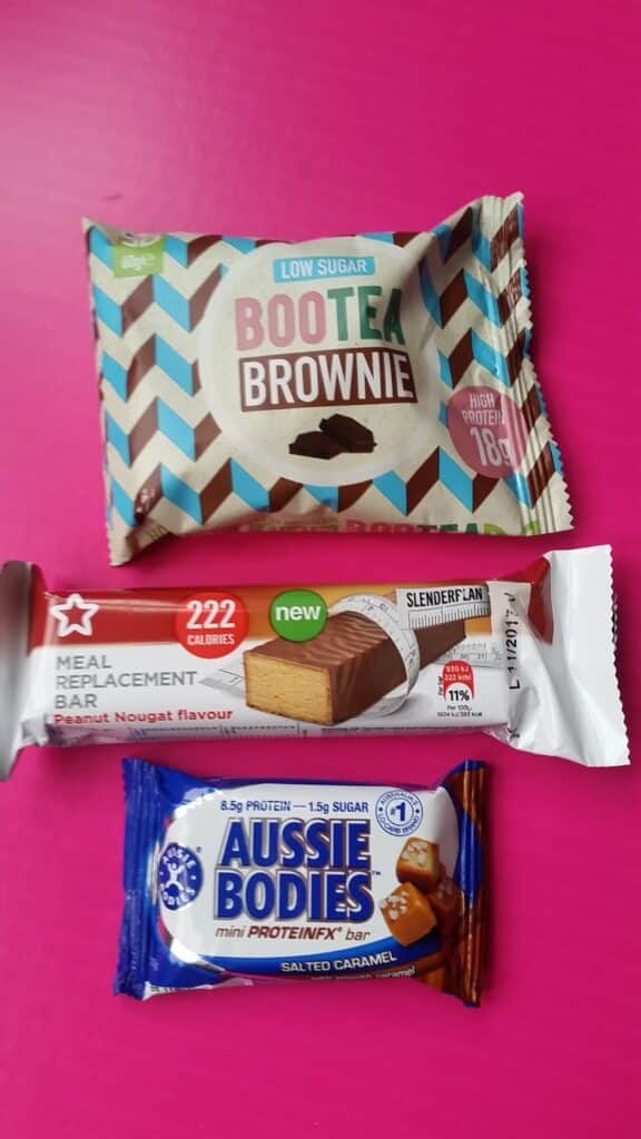 Three protein bars are displayed on a bright pink surface: a BootEA Brownie (blue, brown, white chevron packaging), a Slender Plan Meal Replacement Bar (red and white, peanut nougat flavour), and an Aussie Bodies mini ProteinFX bar (blue, salted caramel).