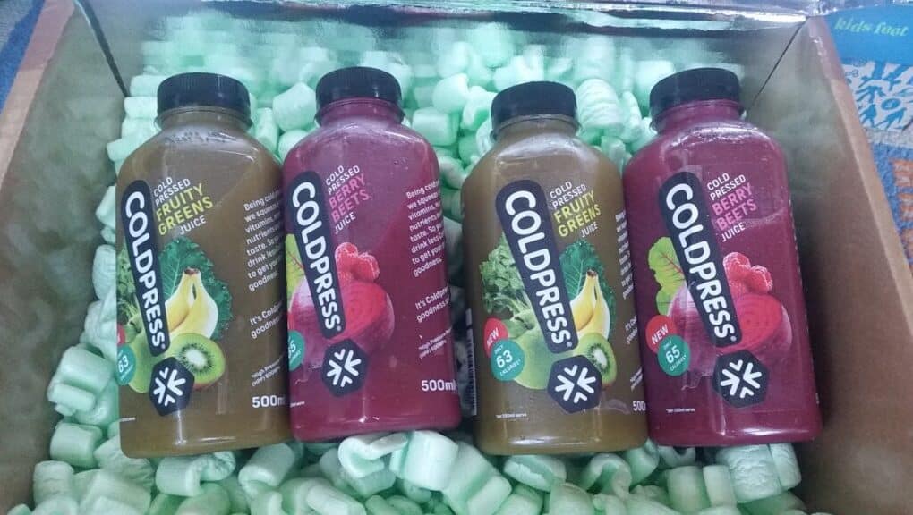 Four Coldpress juice bottles rest in a box with green packing peanuts. Two bottles are labelled “Fruity Greens Juice” (green label with kiwi and banana), and two are “Crushed Berry Juice” (red label with berries). Each bottle holds 500ml.