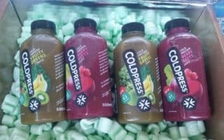Four Coldpress juice bottles rest in a box with green packing peanuts. Two bottles are labelled “Fruity Greens Juice” (green label with kiwi and banana), and two are “Crushed Berry Juice” (red label with berries). Each bottle holds 500ml.